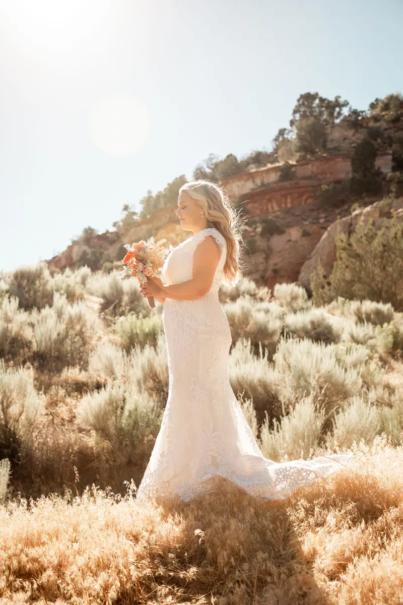 Bride in elegant lace dress holding bouquet in sunlit desert scenery.