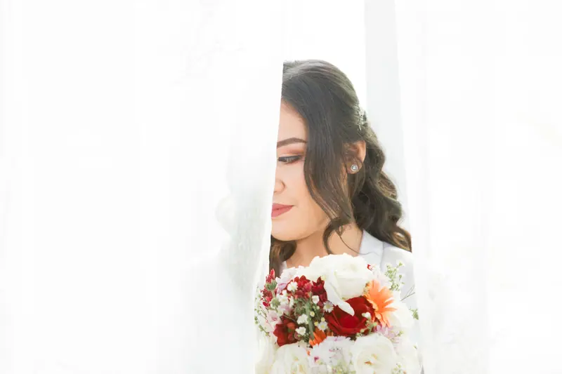 An elegant bride with a floral bouquet thoughtfully concealed behind a curtain, creating a dreamy wedding moment.