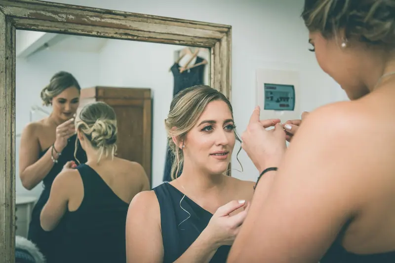 Bridesmaid adjusting jewelry before a wedding, reflected in the mirror, showcasing elegance and friendship.