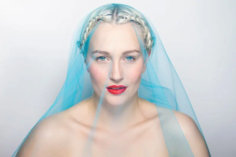 Stunning portrait of a bride with braided hair under a sheer blue veil, highlighting red lips.