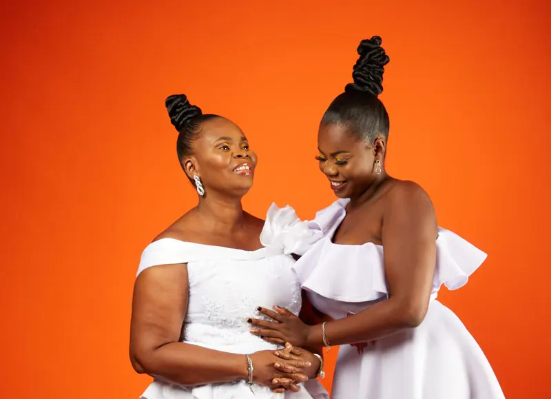 Mother and daughter in elegant white dresses share a joyful moment against an orange backdrop.