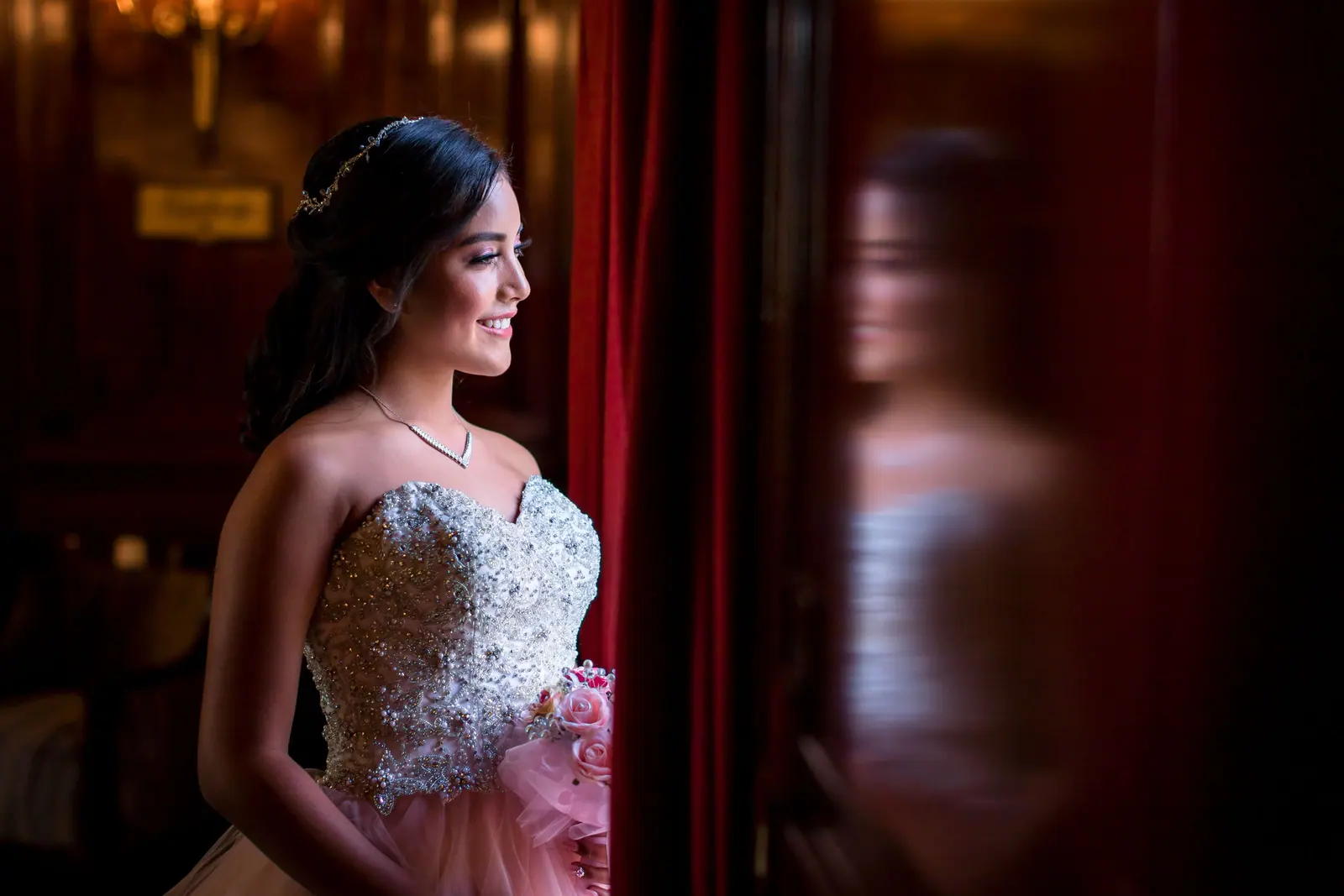 Bride with a sleek updo and tiara in a beaded gown, lit softly against dark red velvet curtains