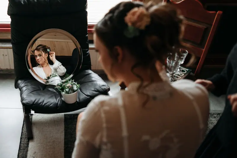 A bride adjusts her hair while looking into a round mirror indoors.