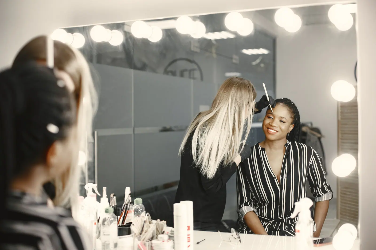 A professional makeup artist applies makeup to a smiling woman in a well-lit backstage setting.
