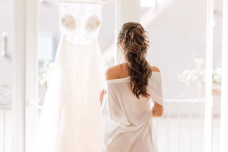 A bride stands with her back turned, gazing at her wedding dress in a serene indoor setting.