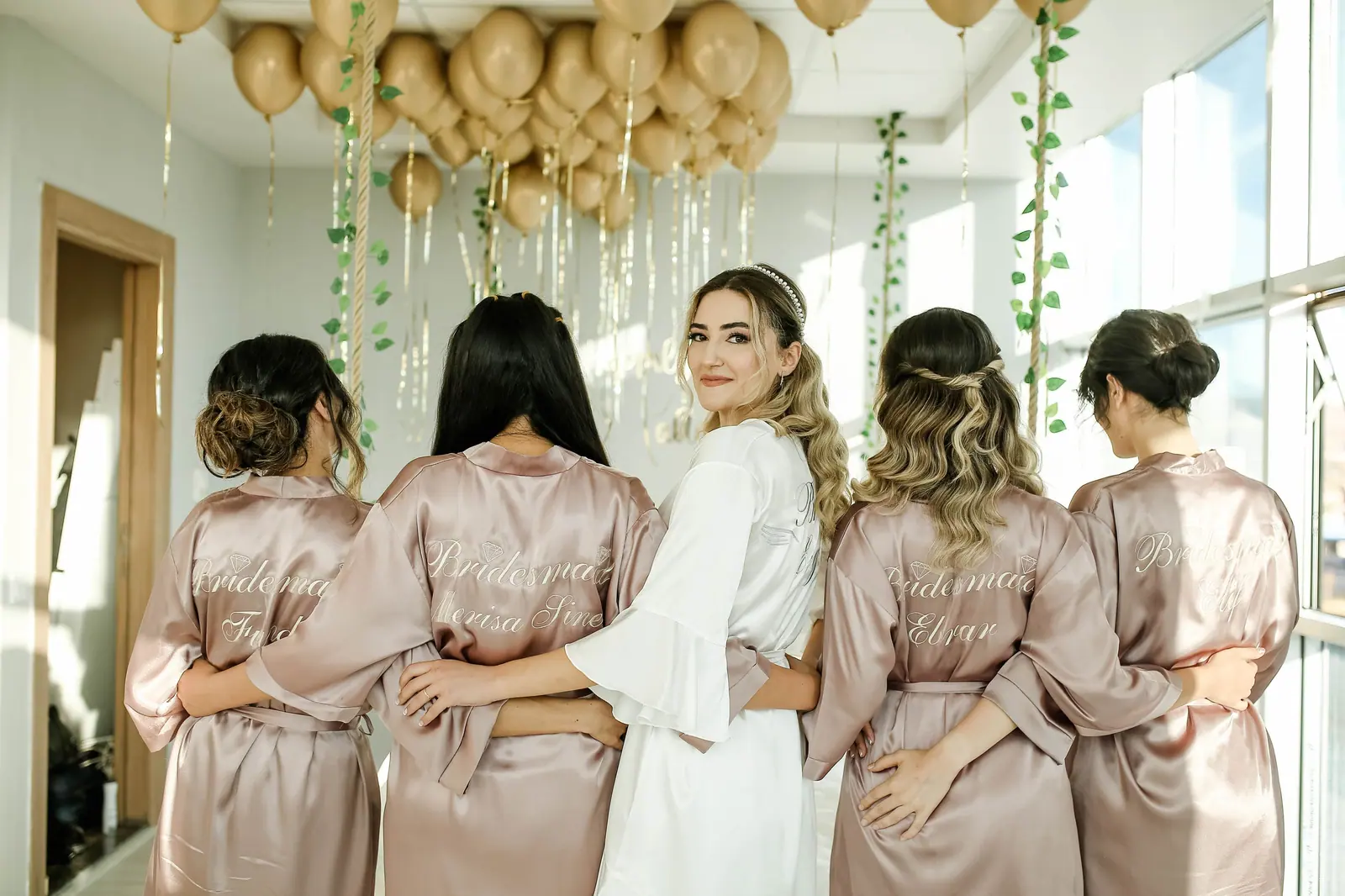 Bride and bridesmaids in champagne satin robes laughing together under gold balloons in a sunlit suite