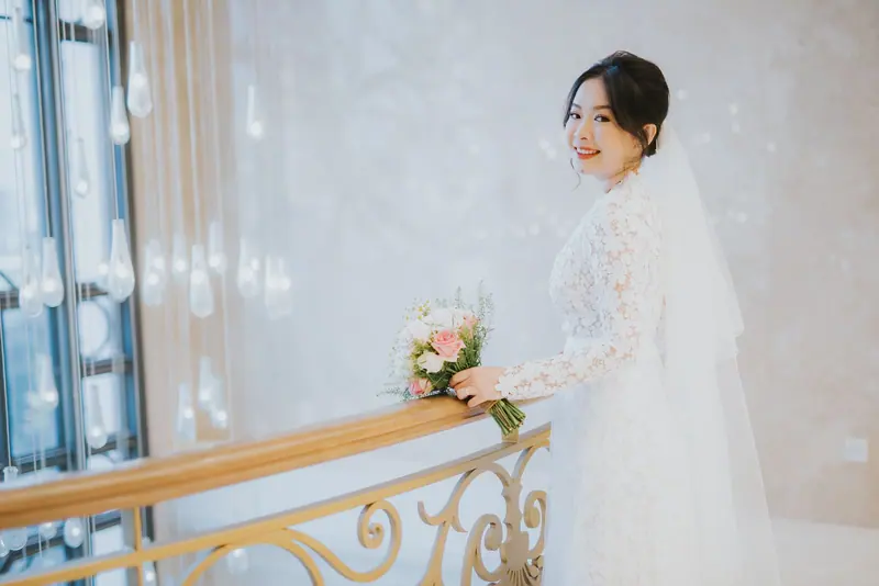 A smiling bride in a lace dress holding a floral bouquet indoors.
