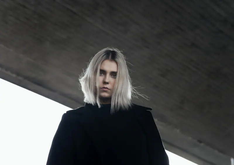 A low angle shot of a serious woman with platinum blonde hair under an urban bridge.