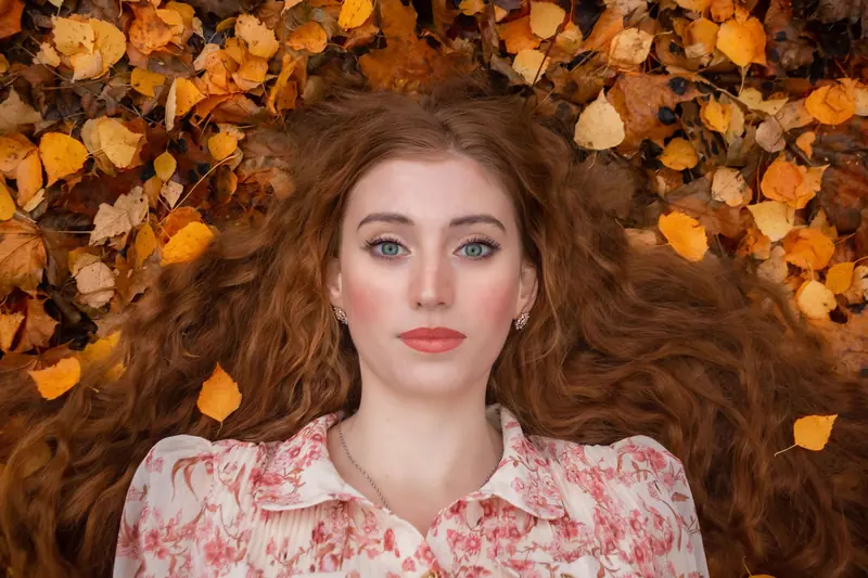Portrait of red-haired woman with makeup among vibrant autumn leaves.
