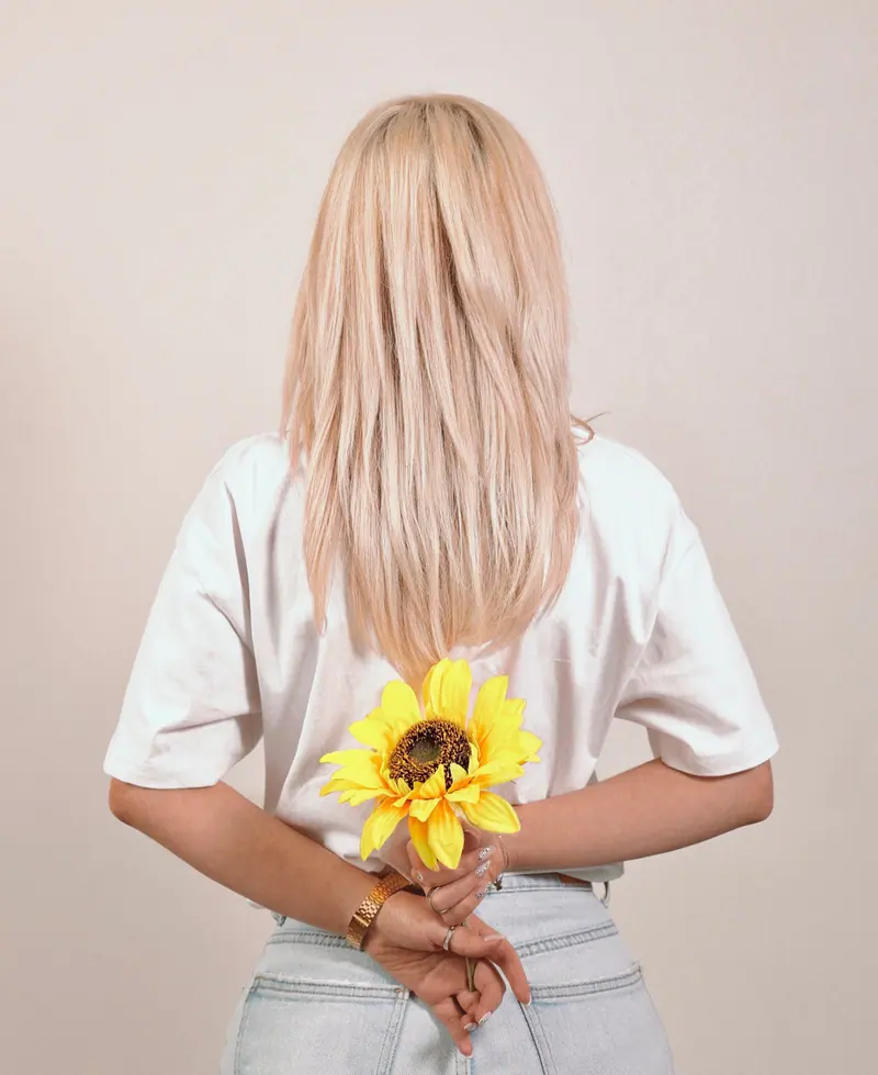 A woman with blond hair holds a sunflower behind her back, captured in an indoor studio setting.