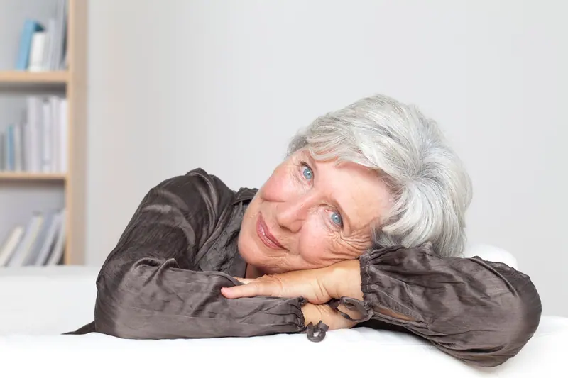 Elderly woman with gray hair smiling and leaning on a table indoors.