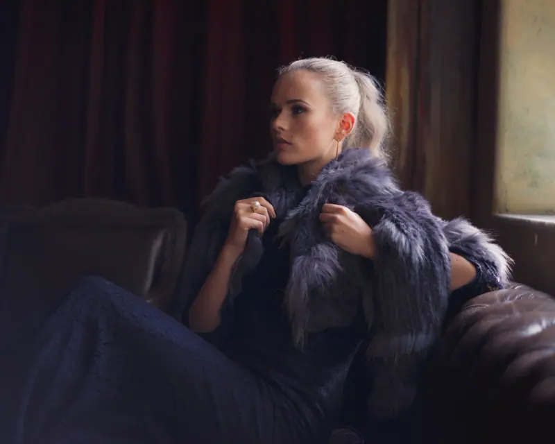 Woman with a sleek blonde ponytail in a violet fur coat on a dark leather sofa