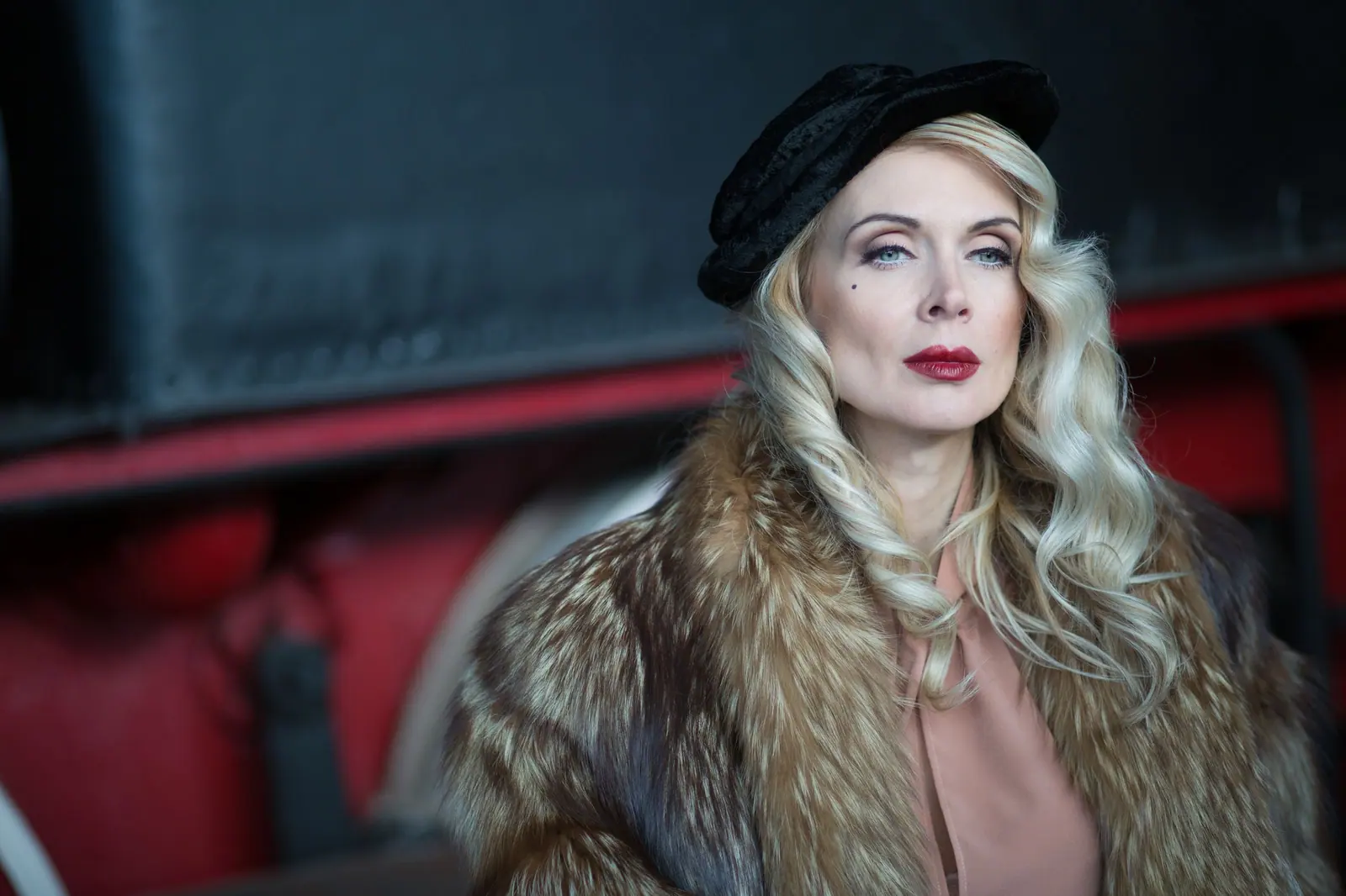 Woman with long platinum waves in a beret and fur collar, lit against a dark red background