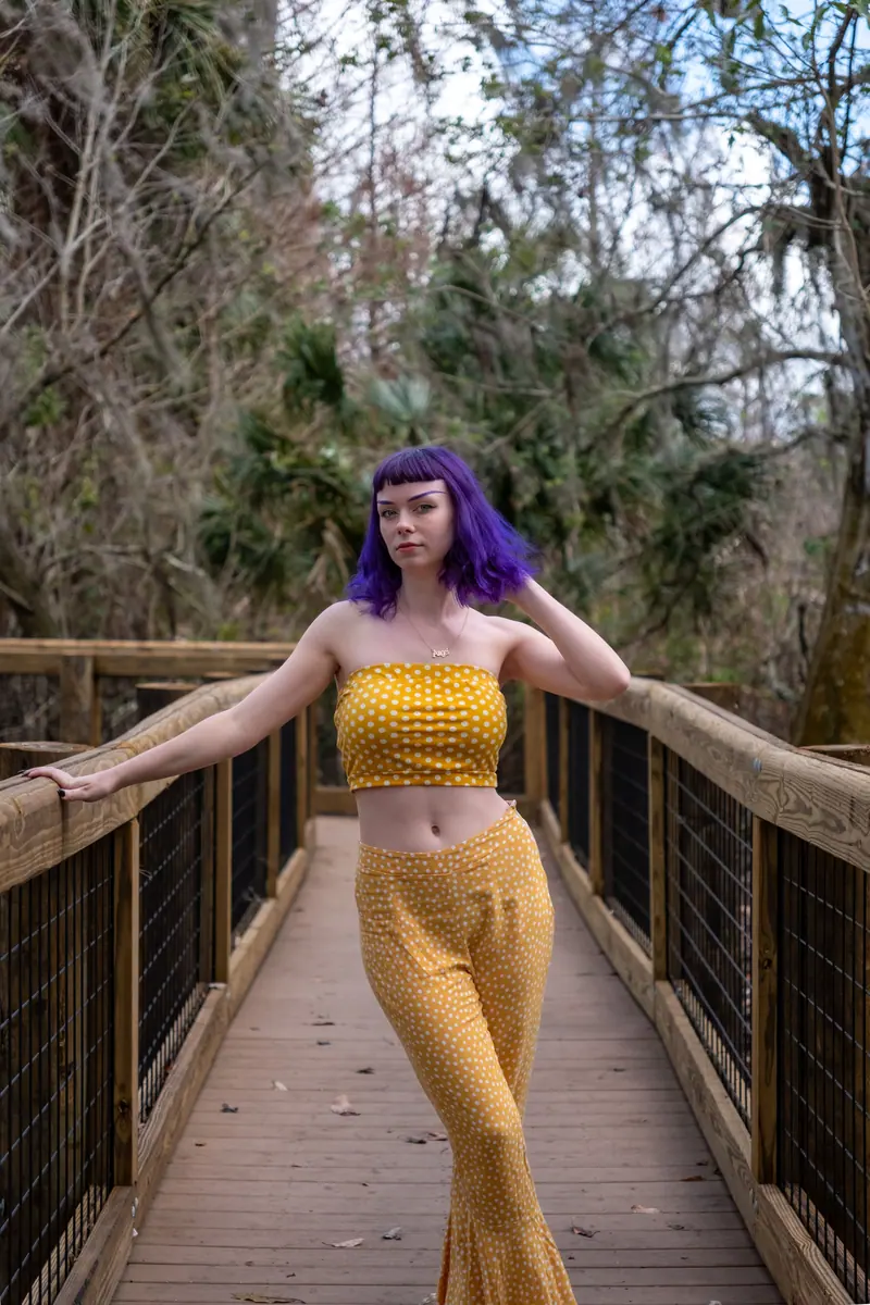 Young woman with purple hair in stylish yellow outfit posing on a wooden walkway in nature.