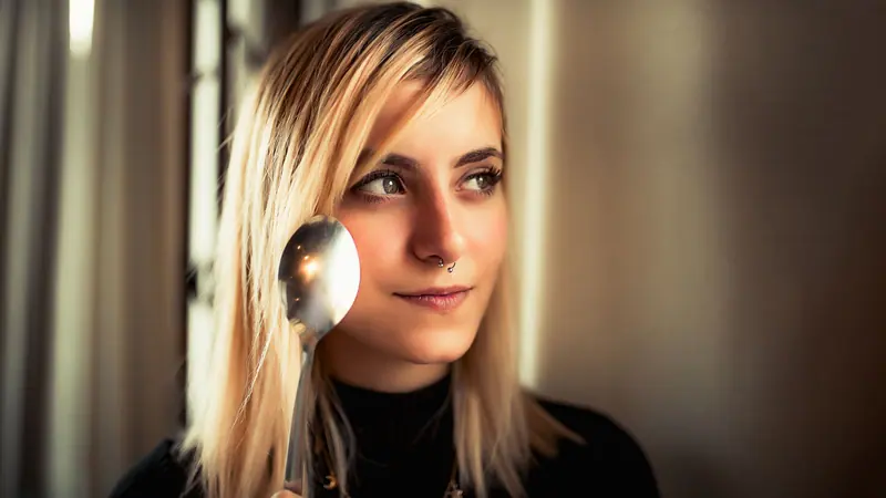 Portrait of a blonde woman with a nose piercing holding a spoon indoors with a soft focus background.