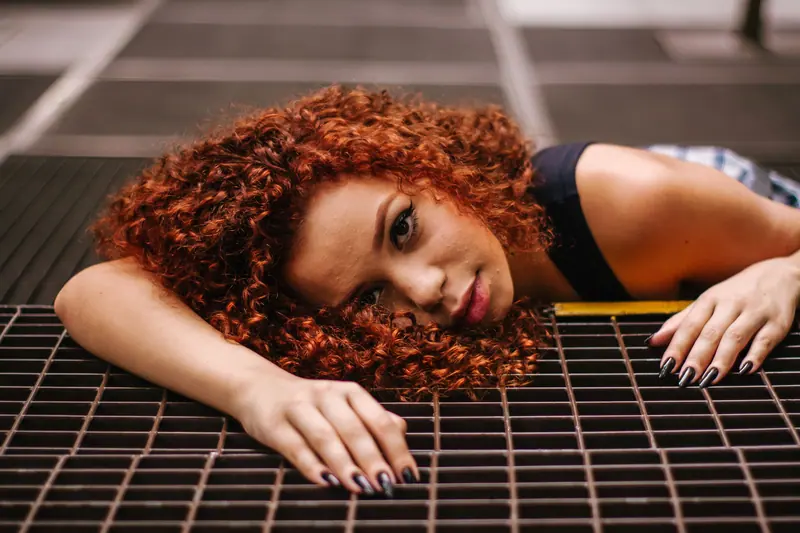 Artistic portrait of a young woman with curly red hair lying on a metal grate in São Paulo.