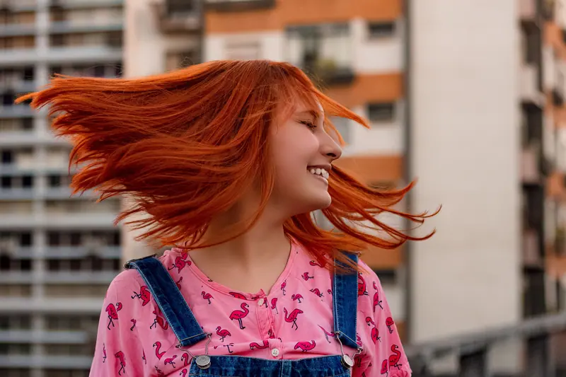 Cheerful woman with vibrant red hair smiling outdoors in urban setting, wearing flamingo print shirt and denim suspenders.