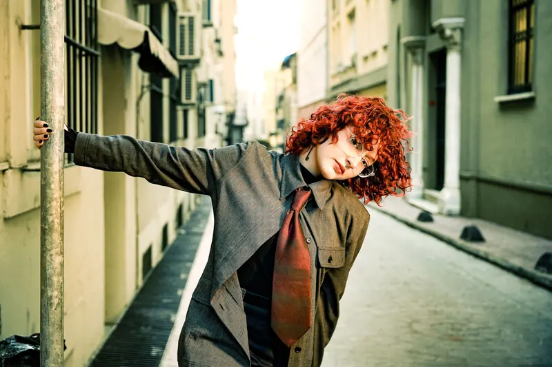 Woman with red curly hair leaning on pole in urban alley. Fashionable and modern city scene.