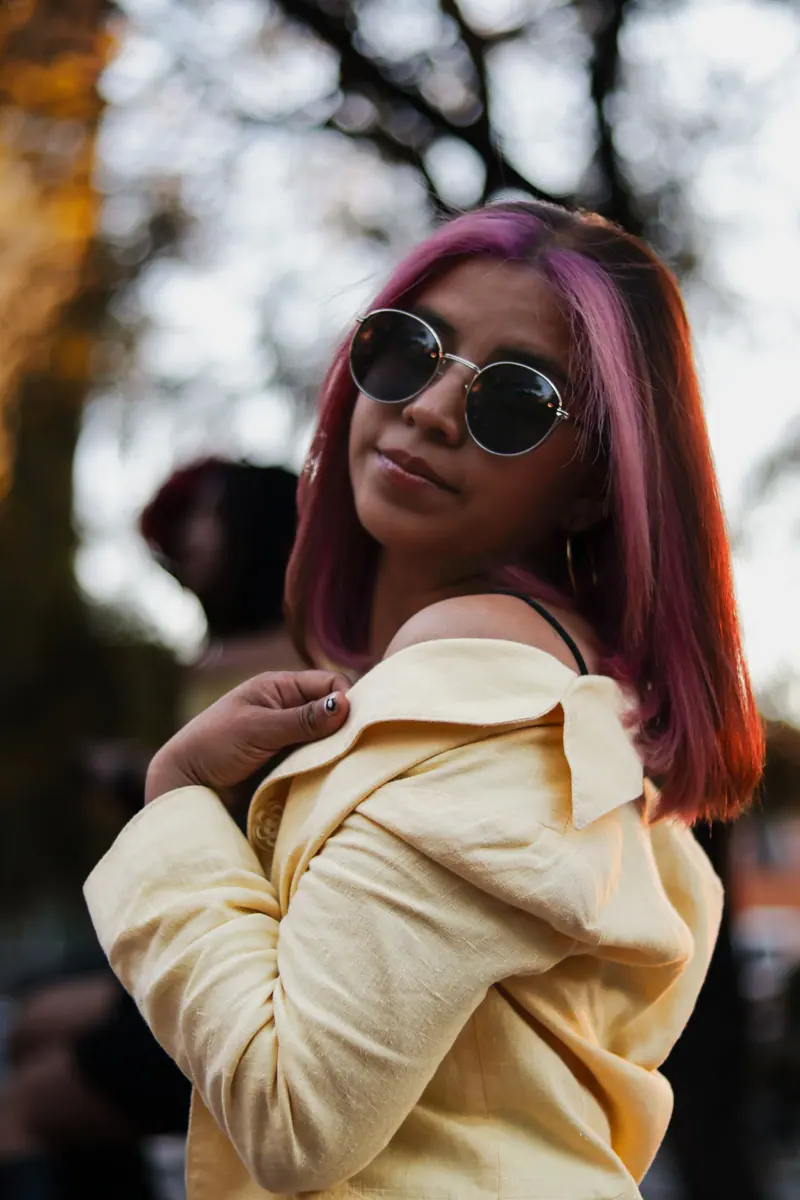 Trendy young woman with pink hair and sunglasses posing confidently outdoors at sunset.