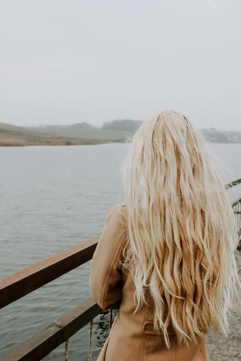 A woman with long blonde hair stands by a railing overlooking a calm lake on an overcast day in Minsk, Belarus.