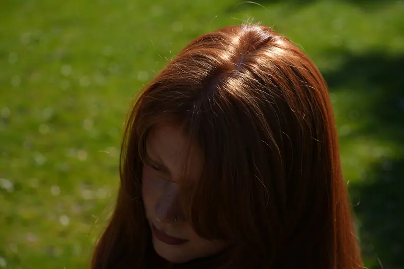 Close-up shot of a woman with red hair in the sunlight, set against a green outdoor background.