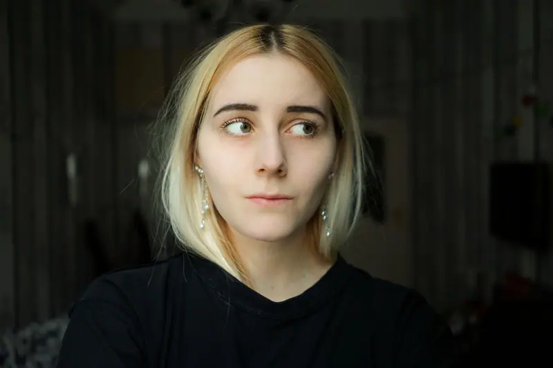 A thoughtful young woman with blond hair looks to the side indoors, reflecting quietly.