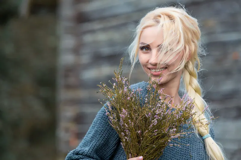 A smiling blonde woman in a knitted sweater holding wildflowers outdoors.