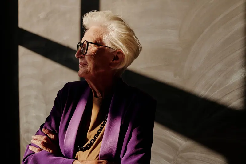 Elderly woman with eyeglasses poses in sunlight casting shadows indoors.