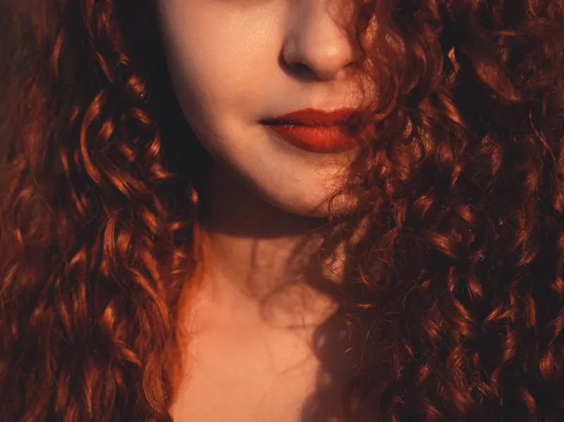 Artistic close-up of a woman with curly red hair and red lipstick in warm sunlight.