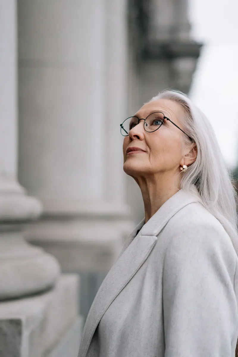 Graceful elderly Caucasian woman wearing eyeglasses looks up thoughtfully outside a historic building.