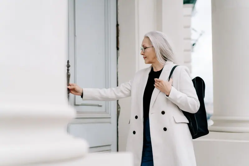 Stylish senior woman in white coat opening a door while standing outdoors, exuding confidence and grace.