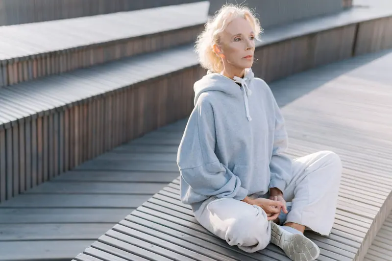 Elderly woman meditating outdoors in sunlight. Relaxation and healthy lifestyle in soft gray sportswear.