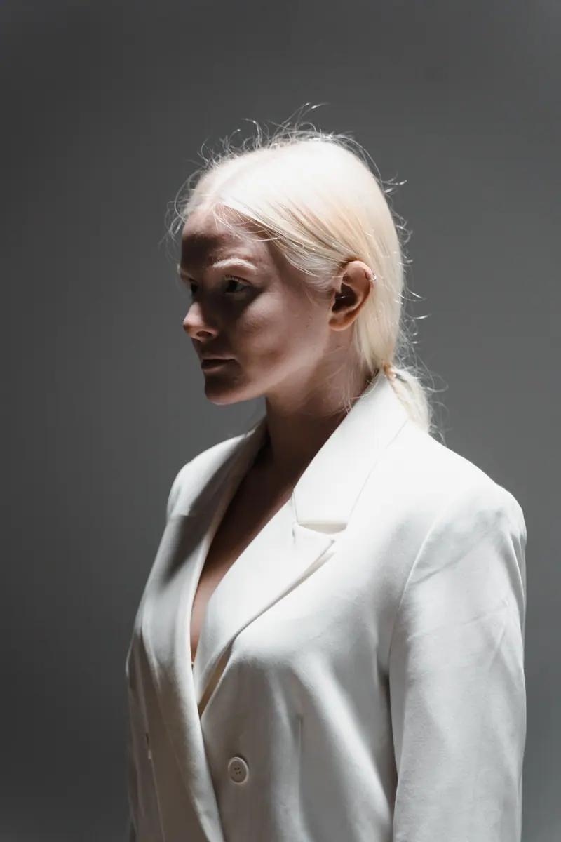 Portrait of a woman in a white blazer with platinum hair against a gray background, in a side view pose.
