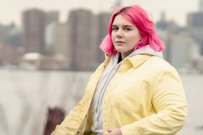 A woman with pink hair stands outdoors wearing a yellow jacket, with a city skyline in the background.