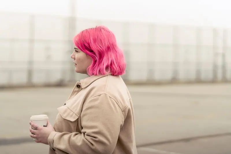A young woman with vibrant pink hair holds a coffee cup outdoors, showcasing individual style and modern urban lifestyle.