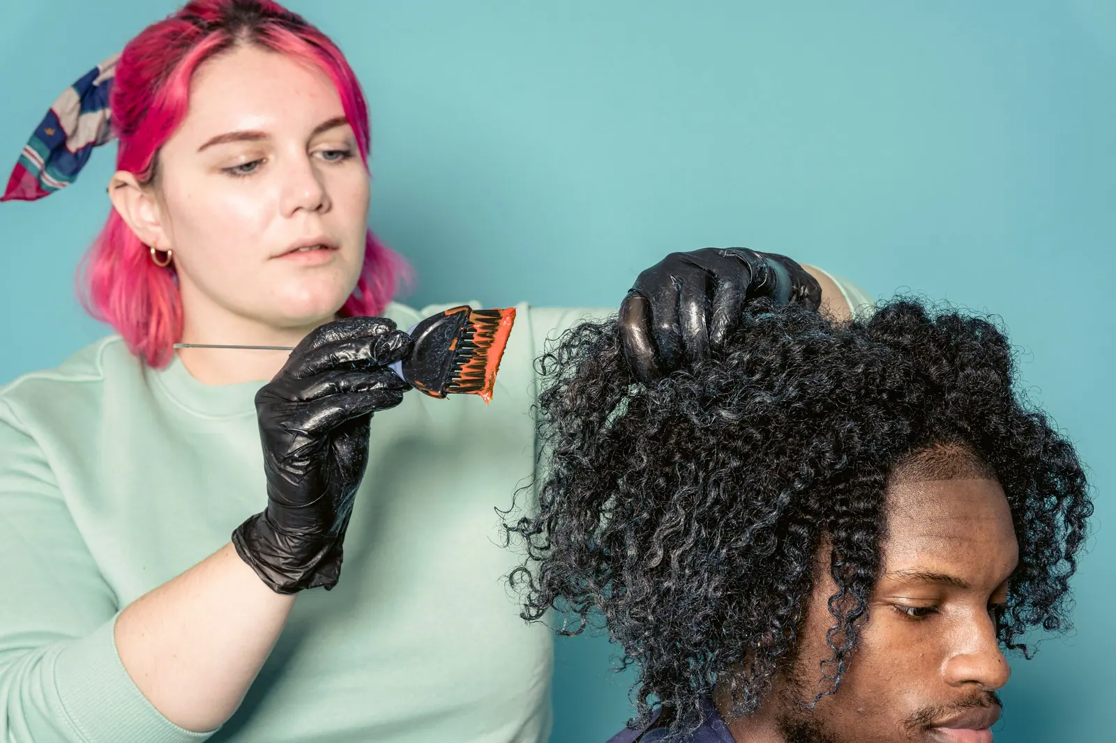 Pink-haired colorist painting color into a client's natural curls against a teal wall