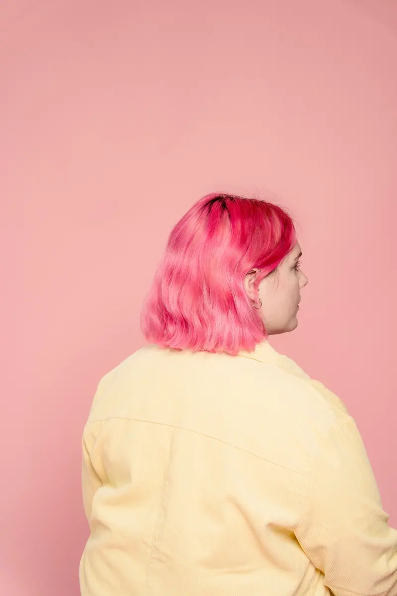 A serene portrait of a woman with pink hair viewed from behind, set against a pastel pink background.