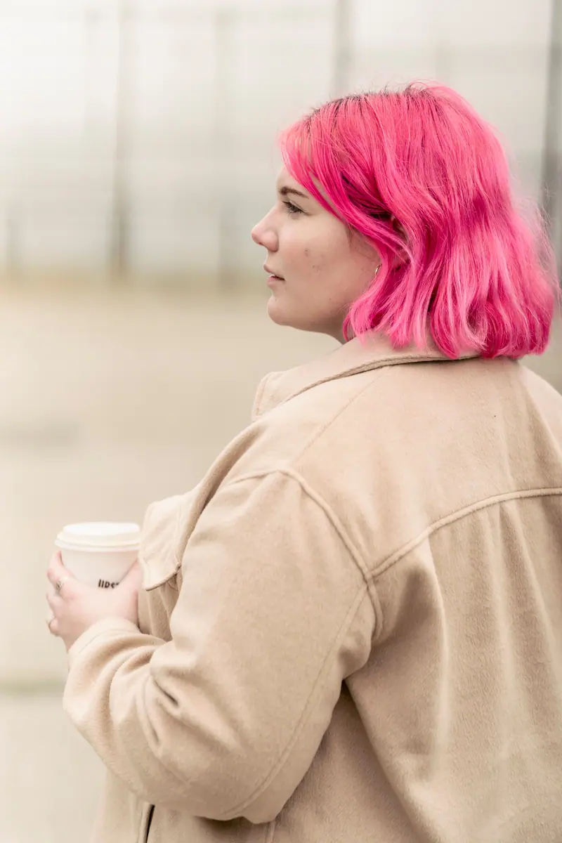 A young woman with pink hair holding a takeaway coffee cup, dressed warmly outdoors.
