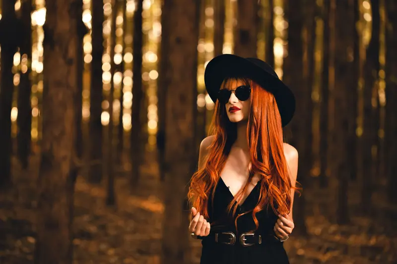 Fashionable woman with red hair in black hat and sunglasses poses in a forest. Elegant style and blurred background enhance the scene.