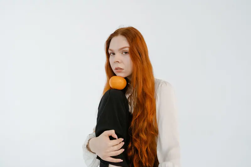 A woman with long red hair poses with an orange against a plain white background.