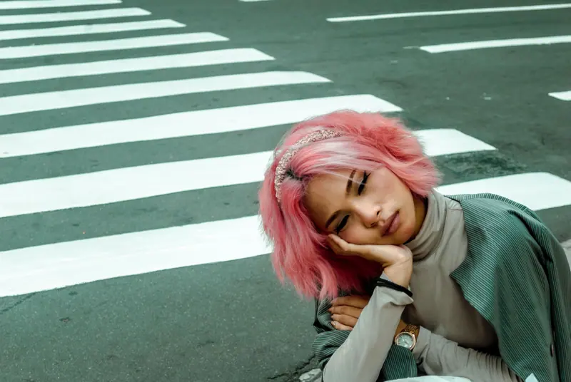 Stylish woman with pink hair and eyes closed, seated at a zebra crossing in daylight.