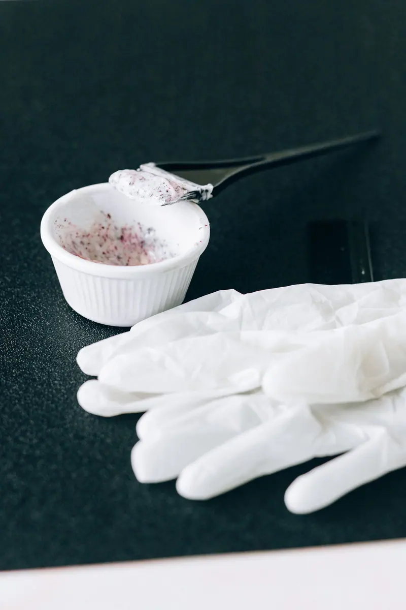 Close-up view of white bowl with hair dye and latex gloves on black surface.