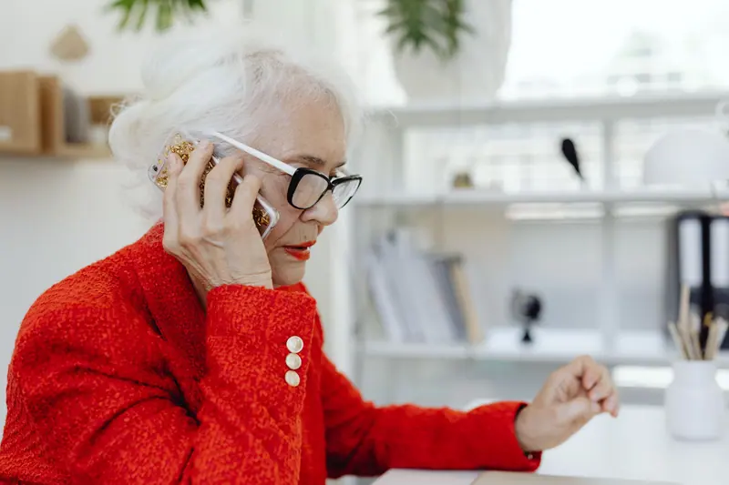 Senior woman in red jacket talking on a smartphone indoors.