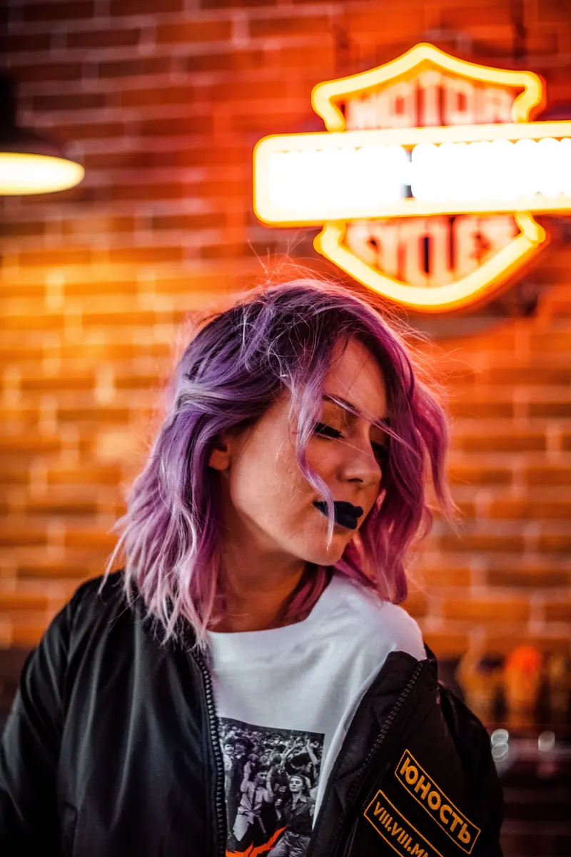 Stylish woman with purple hair and edgy style in front of neon sign in urban area.