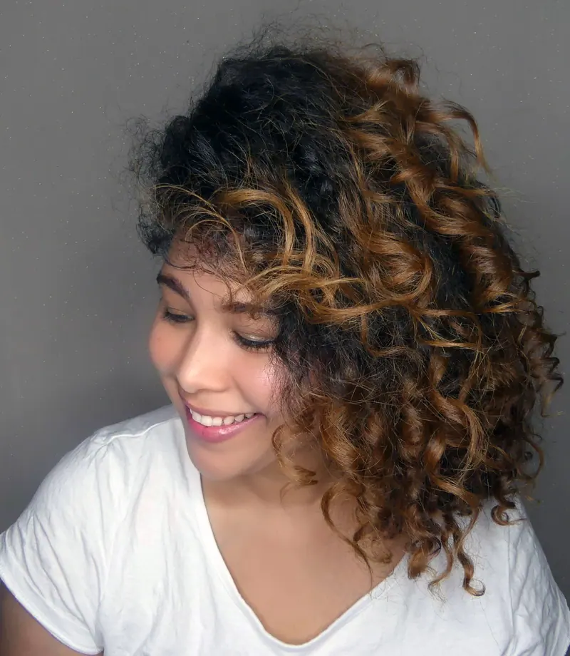 Portrait of a young woman with curly hair smiling indoors against a gray background.