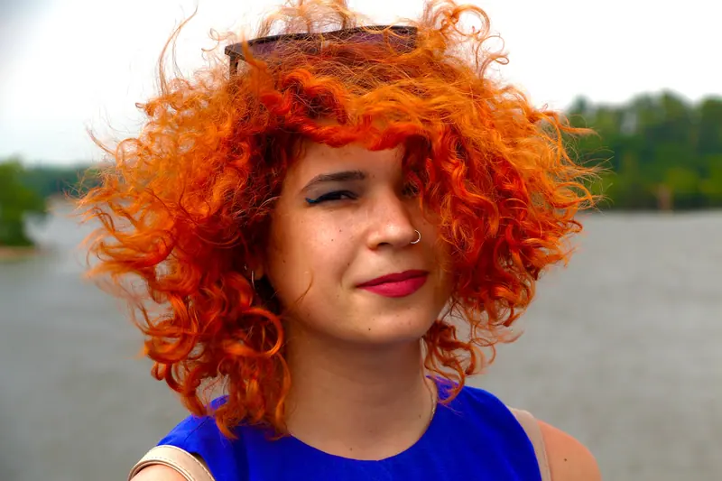 Close-up of a smiling woman with curly red hair and a nose piercing against a natural background.