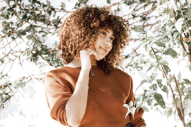 A woman with curly hair in a rust sweater posing outdoors among greenery.