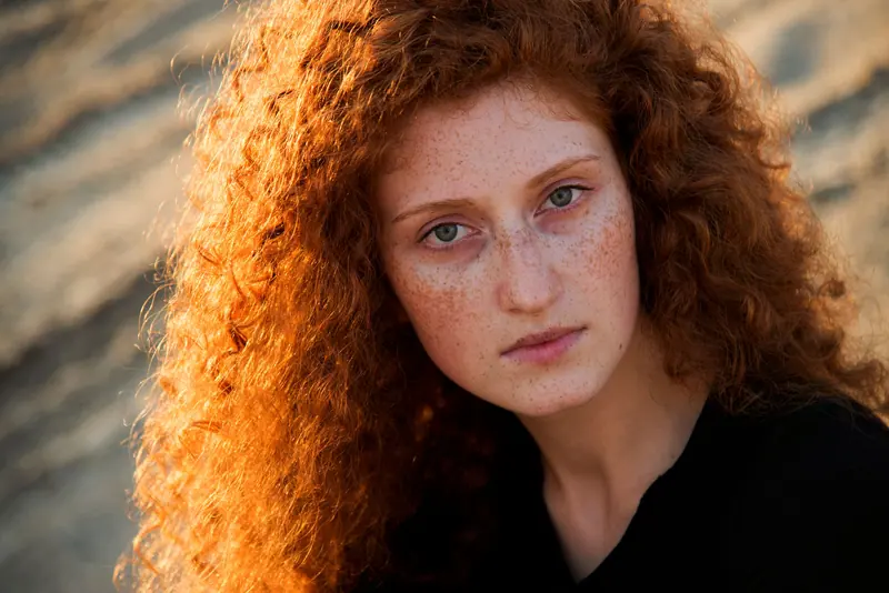 A captivating portrait of a young woman with vibrant red curly hair and freckles in natural sunlight.