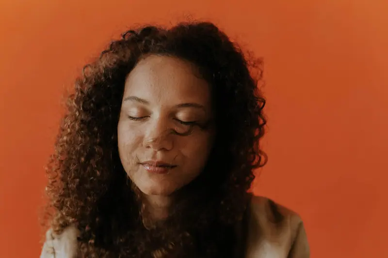 Elegant studio portrait of a woman with curly hair and eyes closed against an orange background.