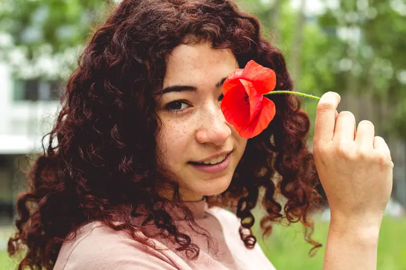 Young woman with curly hair holding a red poppy flower outdoors, smiling softly.